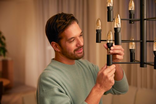 Person installing filament candle bulbs on a black chandelier in a softly lit living room with curtains in the background.