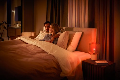Two people sitting together in bed in a softly lit bedroom, with a table lamp emitting red light on the bedside table.
