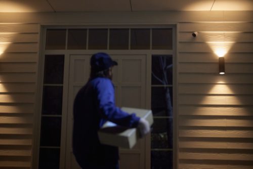 A courier delivers a package at night to a house entrance, illuminated by wall-mounted outdoor lighting and monitored by a security camera.