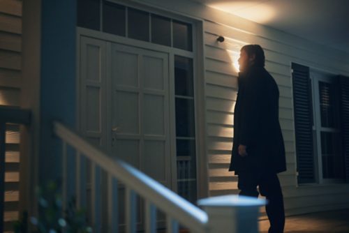 A person stands at a front porch door at night, illuminated by a wall-mounted outdoor security light above the entrance.