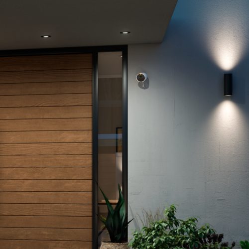 Wired white security camera mounted on exterior wall near door, positioned above plants, with wall light casting illumination below.