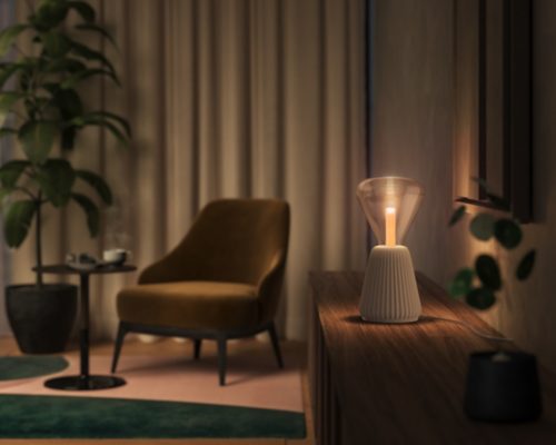 Triangle-shaped glass table lamp with a sand-colored base placed on a wooden surface, emitting soft light onto the wall.